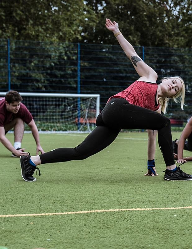 A group class, with an athletic lady stretching and looking at the camera. The class is outside on a 5-a-side football pitch.